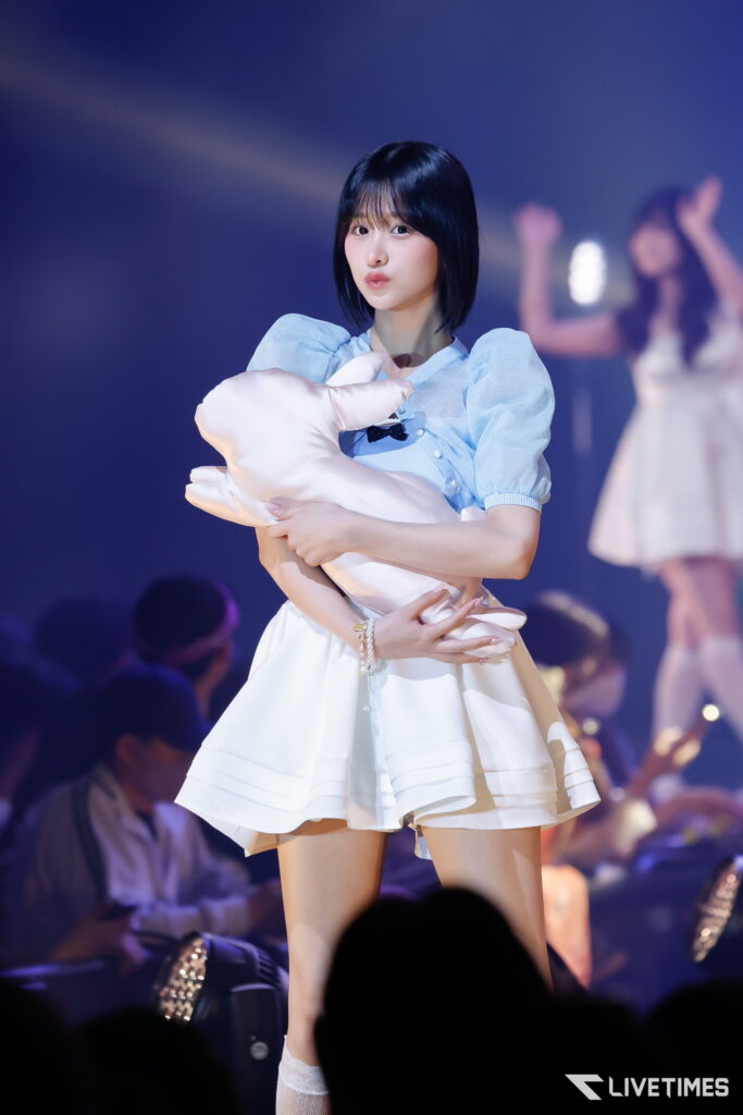 Young female idol in blue blouse on stage, cuddling a pale pink plush pillow amid blue spotlight and cheering crowd.