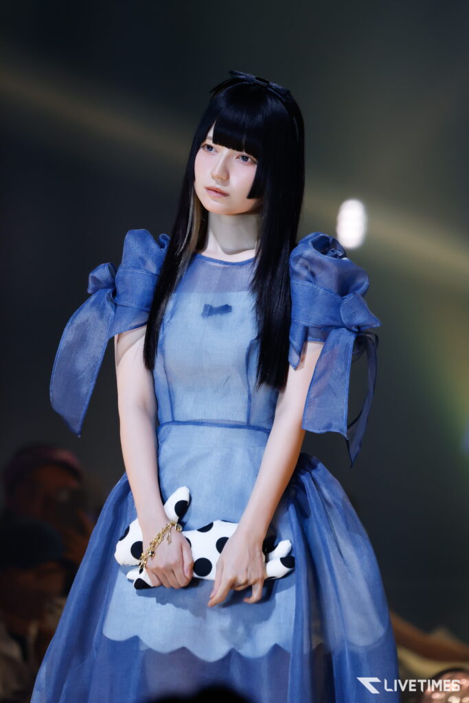 Young woman in a blue dress with puff sleeves on a runway, holding a white polka-dot clutch.