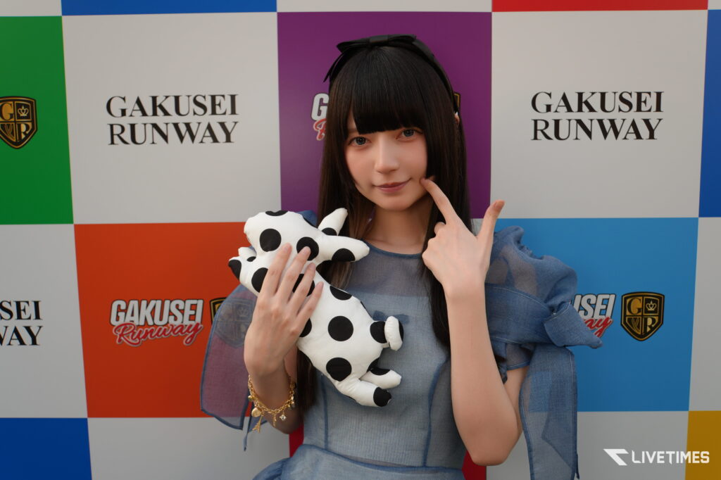 Young woman posing in front of a colorful Gakusei Runway backdrop, holding a black-and-white polka-dot plush toy.