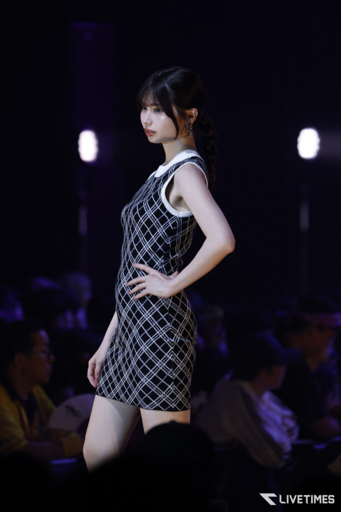 Female model walks the runway in a sleeveless black-and-white plaid dress, hands on her hips, spotlighted against a dark background with an audience in silhouette.