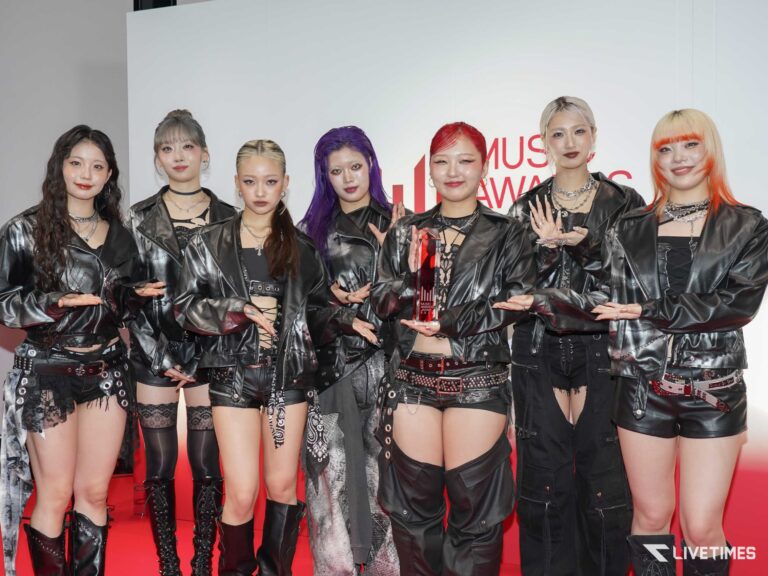 Eight women in black leather outfits pose on a red carpet at a music awards event, one holding a glass trophy with a white backdrop behind them.