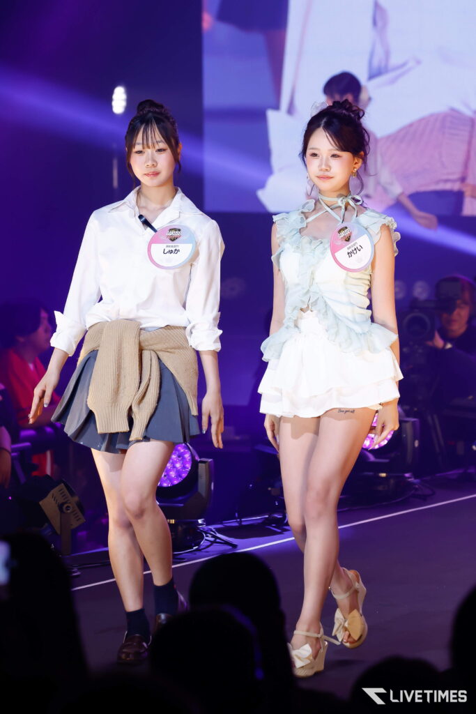 Two young women walk on a lit stage wearing stylish outfits with round name badges pinned to their chests, one in a white blouse and gray skirt, the other in a pale ruffled dress.
