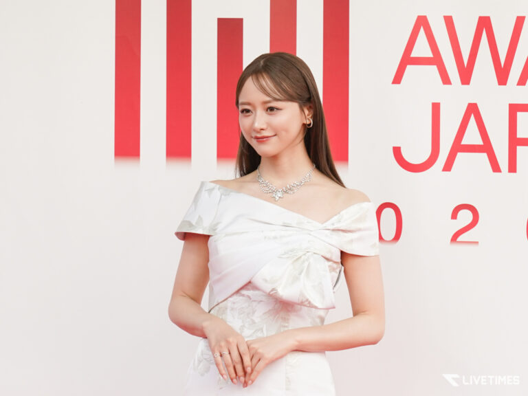 Woman in an off-shoulder white gown with a diamond necklace posing on a red carpet backdrop; LIVETIMES watermark bottom right.