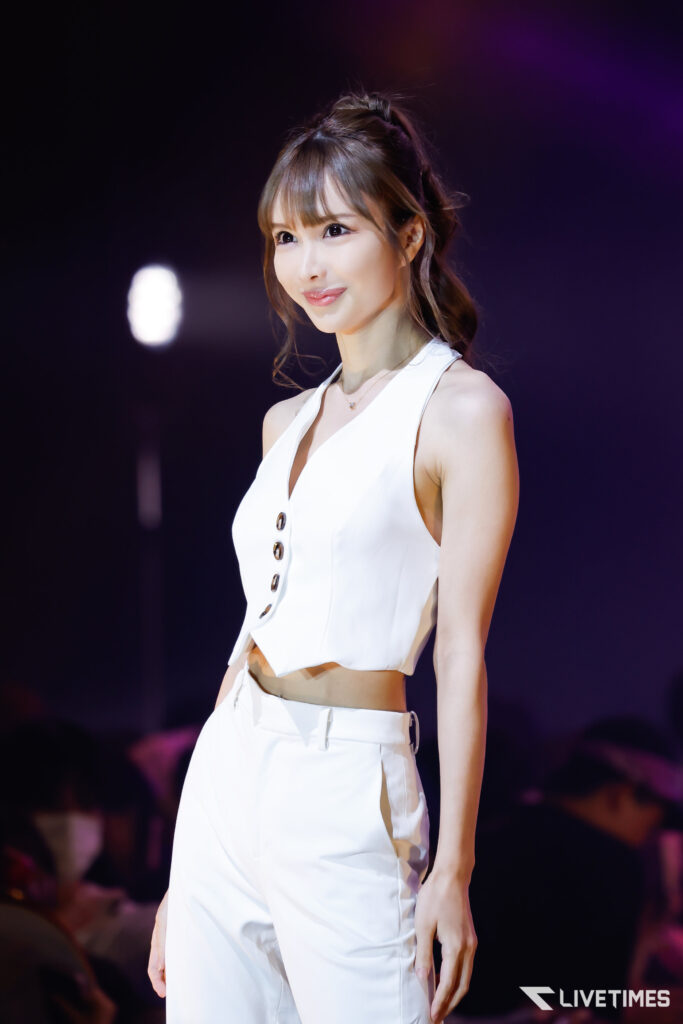 A female performer in a white sleeveless outfit standing on stage with a dark background and purple lighting, smiling subtly at the audience.