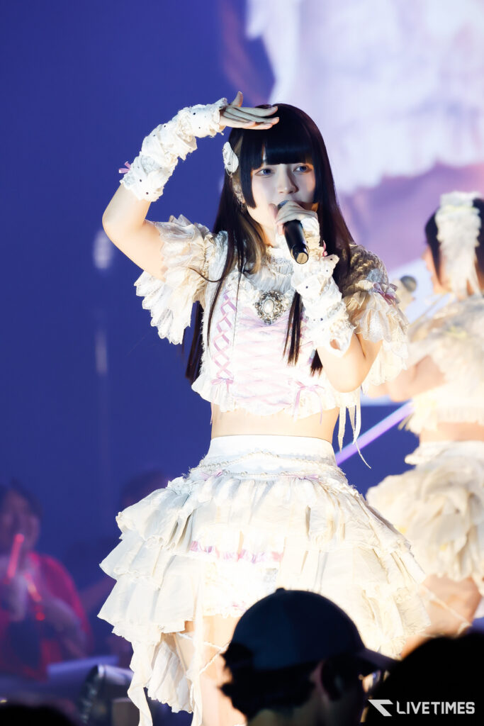Idol singer in a white frilly costume performs on stage, shielding her eyes from lights with one hand while holding a microphone with the other.