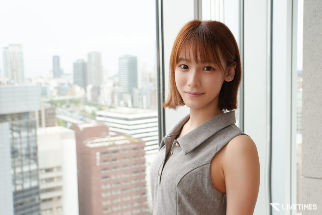 Portrait of a woman with short brown hair standing by a window overlooking a city skyline.]