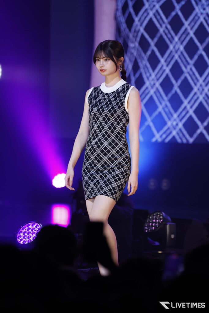 Female performer on stage wearing a black-and-white plaid sleeveless dress under purple stage lights, audience in foreground.