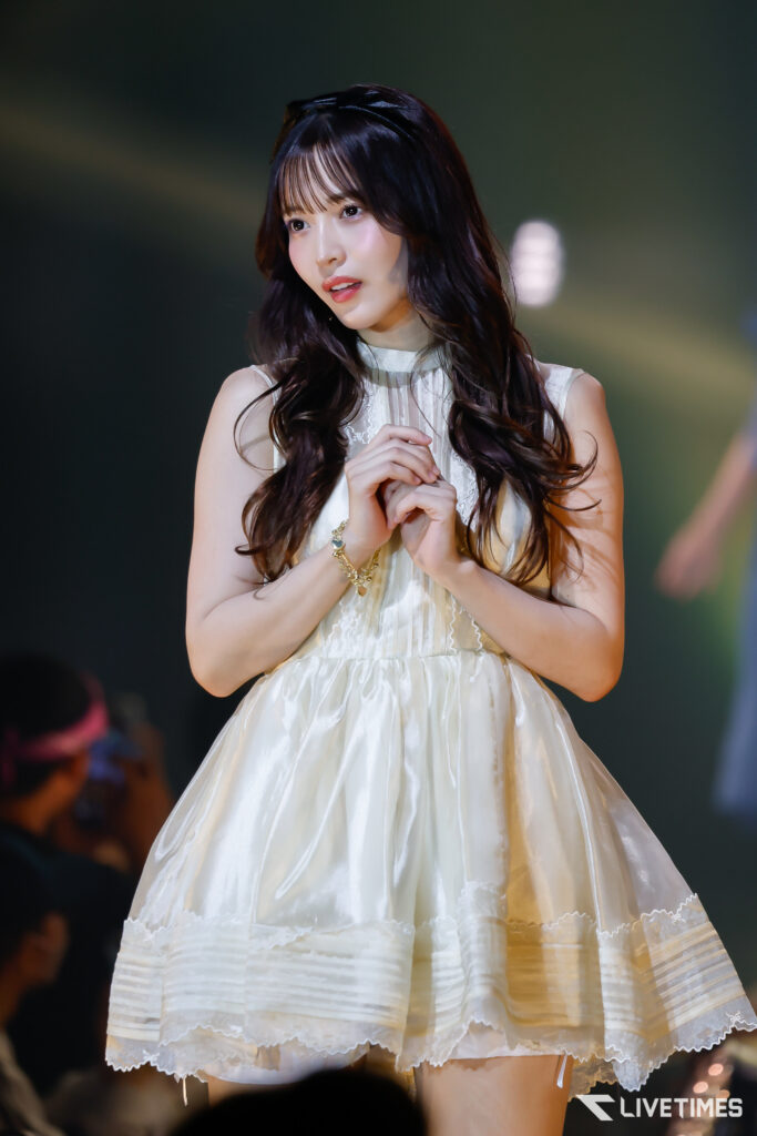 Female singer on stage wearing a white, lacy dress with hands clasped in front, under spotlight.