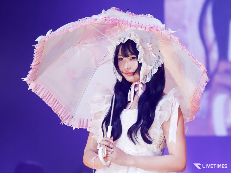 Young woman in a pastel pink Lolita dress holding a decorative parasol on stage, smiling at the audience.