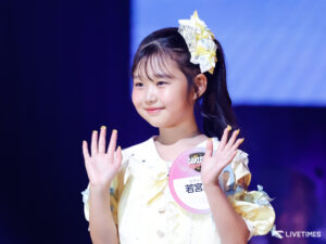Young girl performing on stage, hands raised, wearing a pale yellow and white outfit with a decorative bow in her hair.