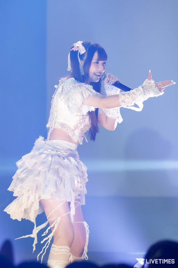Female idol performing on stage in a white frilly outfit, holding a microphone and smiling at the crowd.