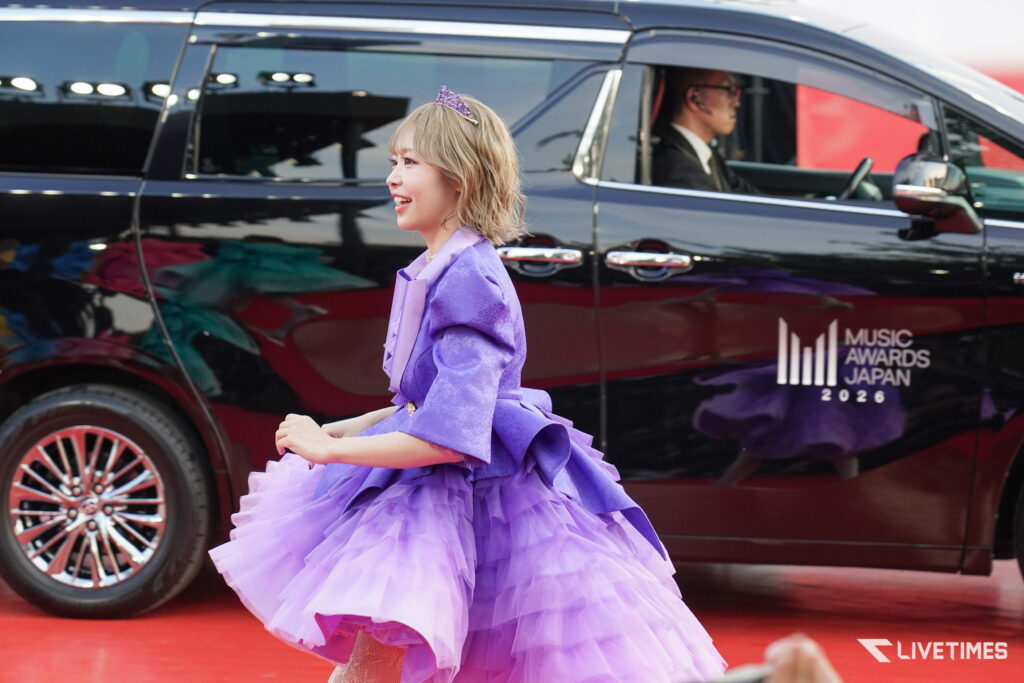 Woman in a lavender gown with a tiara walking on a red carpet near a black event van (Music Awards Japan 2026).