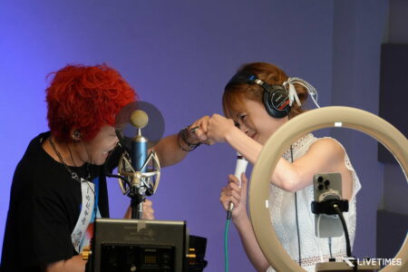 Two people in a radio studio adjusting a microphone and equipment together, with a blue background.