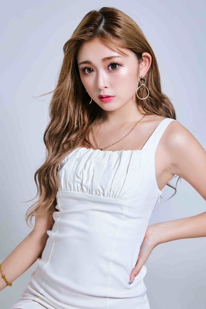 Woman with long wavy hair in a white off-shoulder dress, wearing gold hoop earrings and a delicate chain necklace, posing with hand on hip against a light gray backdrop.