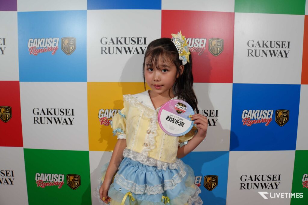 Young girl in a pastel yellow and blue dress posing on a colorful checkered backdrop with 'Gakusei Runway' logos, holding a round sign.