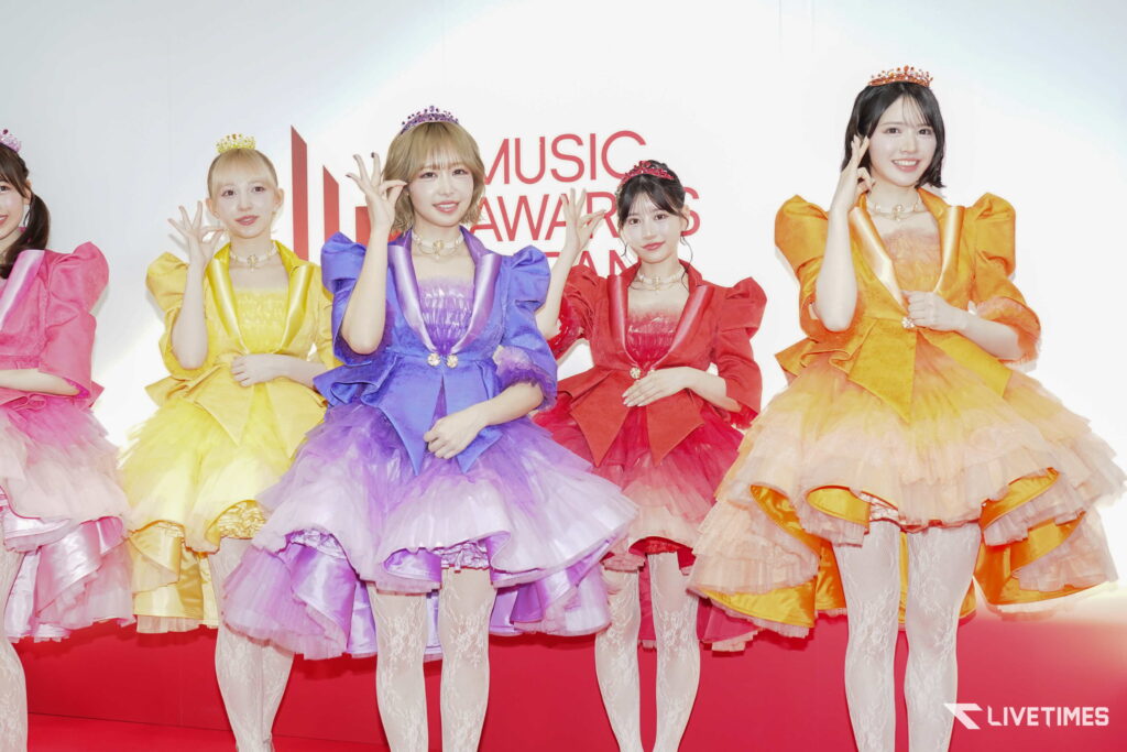 Five women in bright, layered gowns and tiaras pose on a red stage at a music awards event.