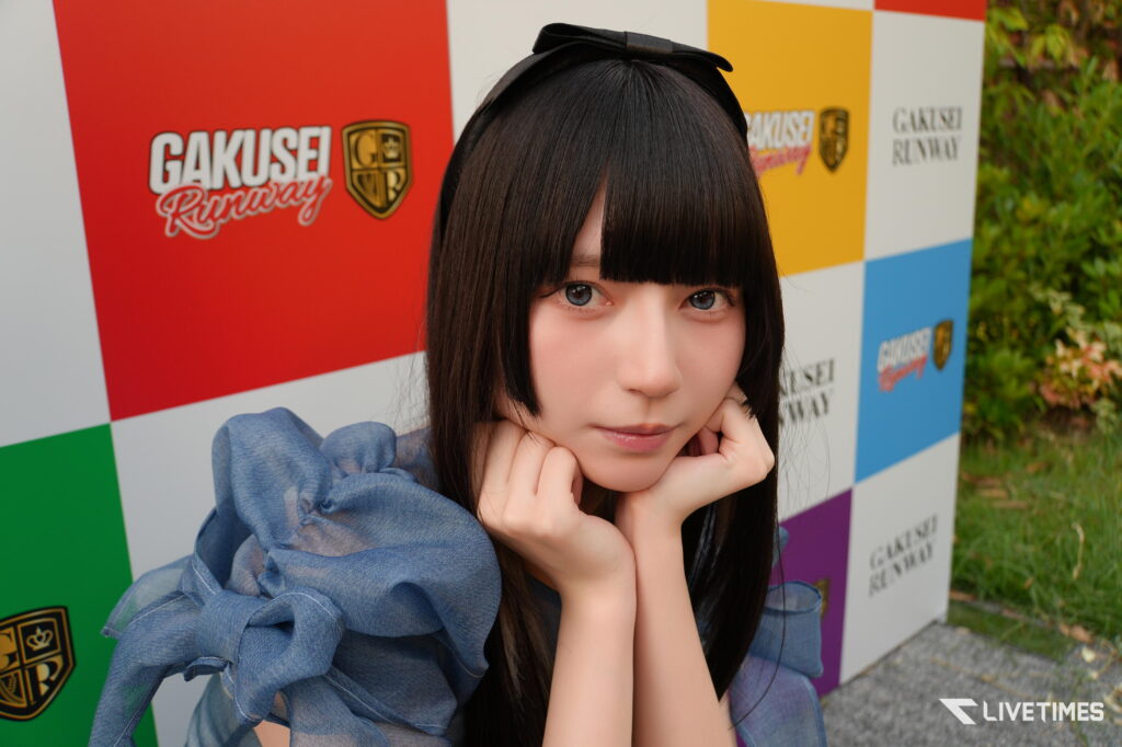 Young woman with straight black hair and bangs posing with her hands under her chin in front of a red, yellow, and blue backdrop with 'Gakusei Runway' logos.