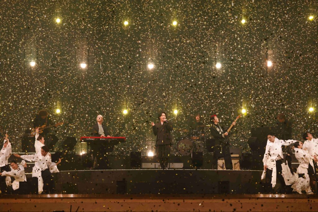 Live concert on a lit stage as gold confetti falls around a band performing and dancers in white outfits fill the foreground.
