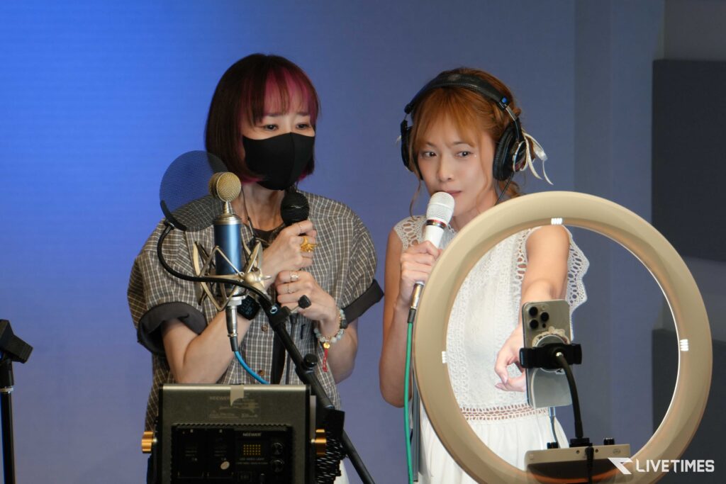 Two women recording a podcast in a studio, speaking into handheld microphones while wearing headsets and a ring light nearby.