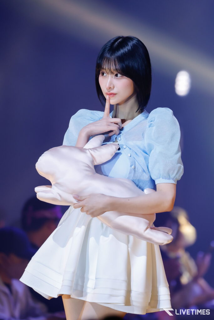 Woman on stage in a light blue blouse holding a white pillow, finger to lips in a shushing gesture with blurred audience in the background (theatre performance).