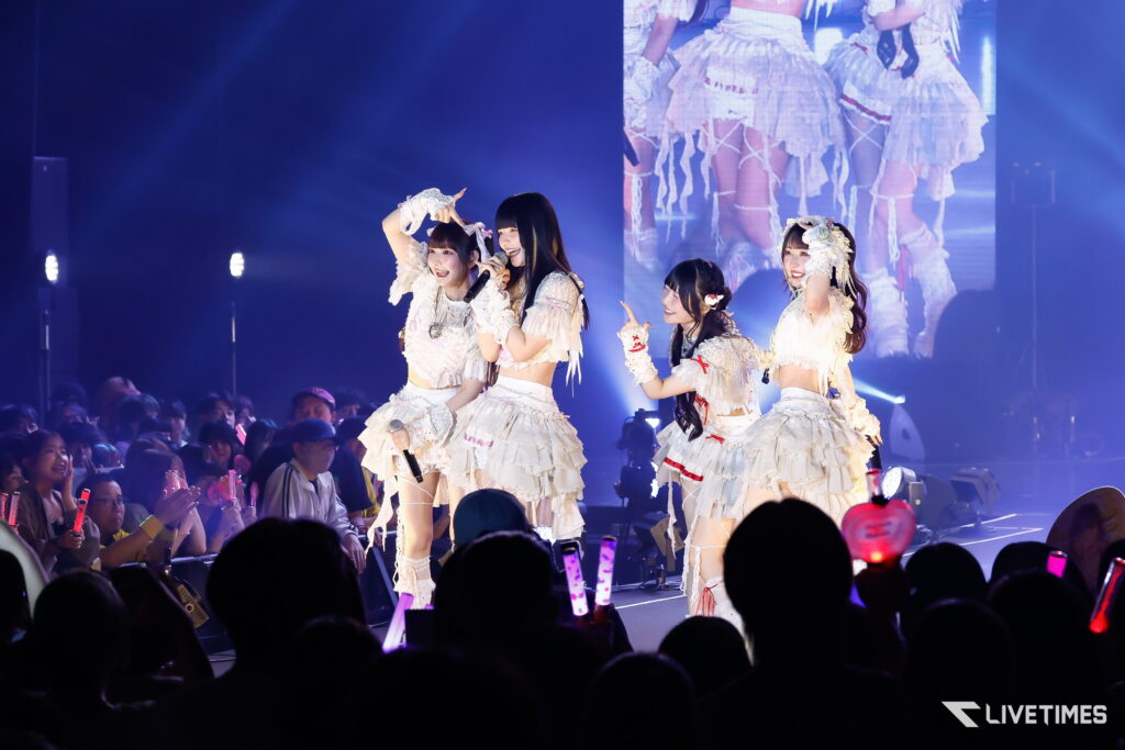 Four female performers in white frilly outfits sing on stage as a crowd cheers under blue lights.