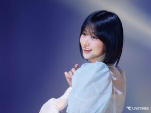 Young woman on stage smiling while looking to the side against a blue gradient background.