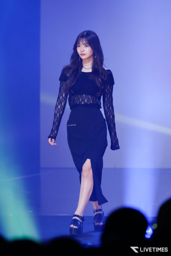 Model walking on a dimly lit runway in a black lace top and high-slit skirt, surrounded by blue stage lighting.