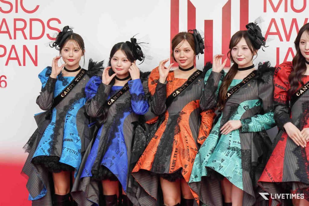 Group of young women in colorful, patterned dresses posing on a red carpet at an awards show with black lace details and headpieces.