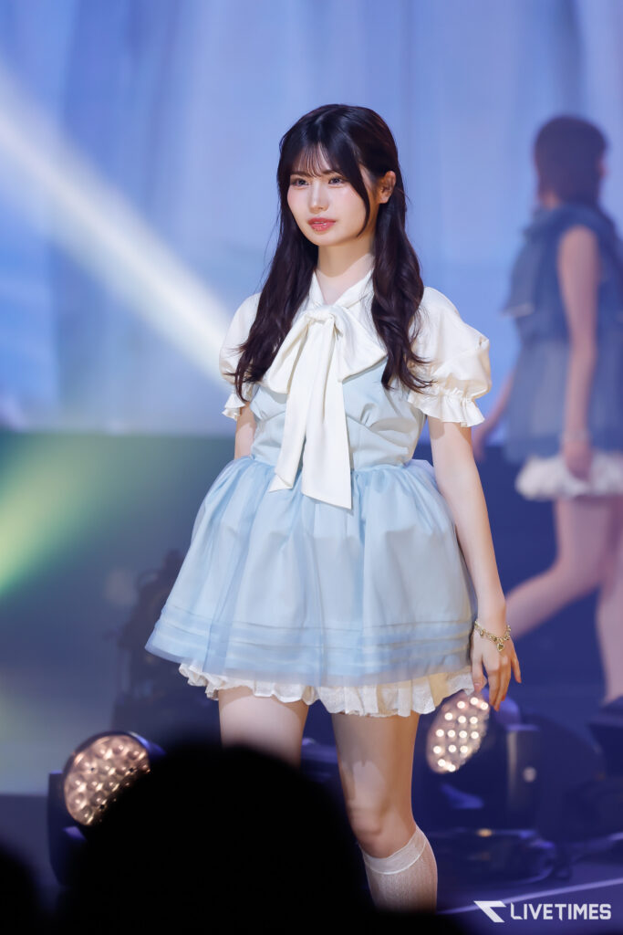 Young woman in a pale blue dress with a white bow performing on a stage under blue lights.