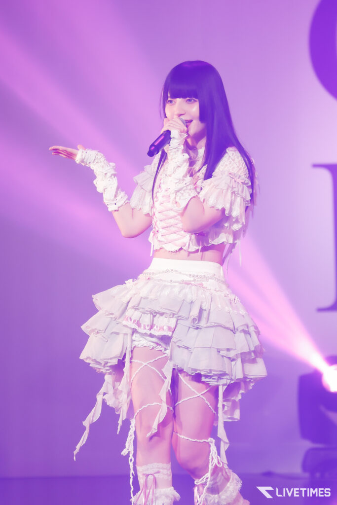 Female singer on stage wearing a ruffled white costume, microphone in hand, performing under pink-purple lights.
