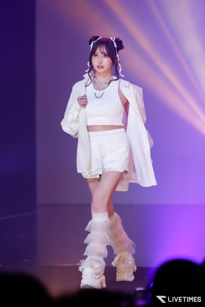 Performer in white outfit with twin buns and ribbon accessories on a purple-lit stage, mid-performance.