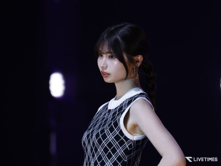 Profile of a female performer on a dark stage, wearing a black-and-white diamond-patterned sleeveless dress and braided hair, looking off to the left.