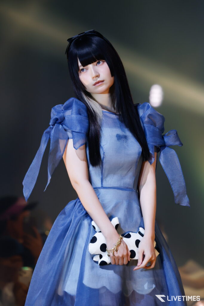 Young woman on stage wearing a blue dress with puff sleeves, holding a white polka-dot clutch and wearing a black wig with bangs.