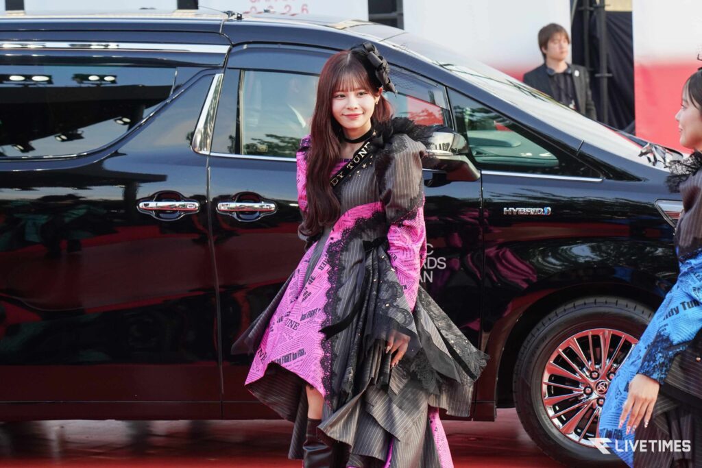 Woman in a pink and black patterned dress posing beside a glossy black SUV at an event.