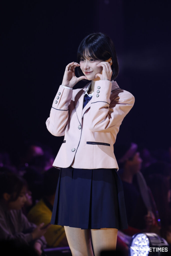 Young woman on stage wearing a pale pink blazer and navy skirt, making a heart shape with her hands toward her face.