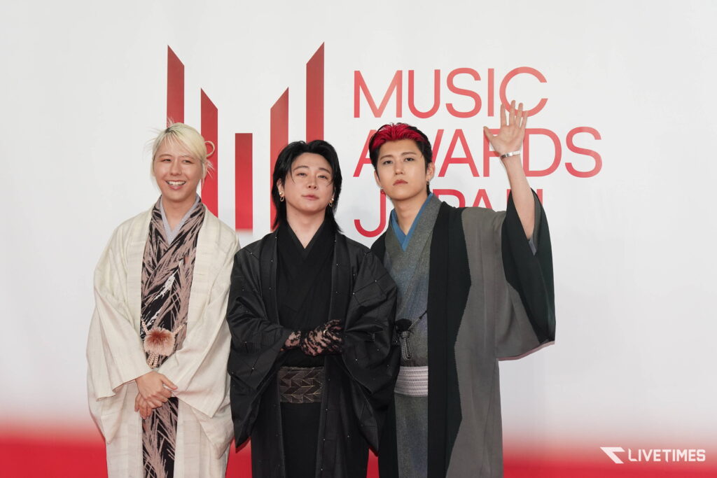 Three people in traditional outfits pose for a photo on a red carpet, with a white backdrop and red event signage behind them.