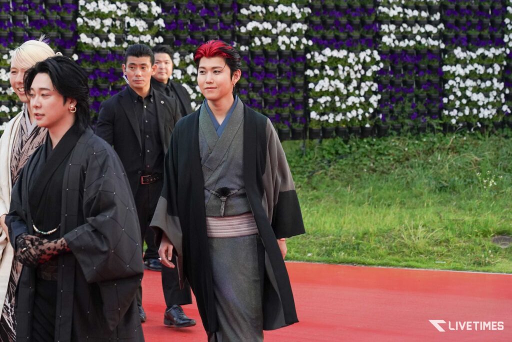 Group of people in dark traditional robes walking on a red carpet beside a wall of purple and white flowers outdoors.