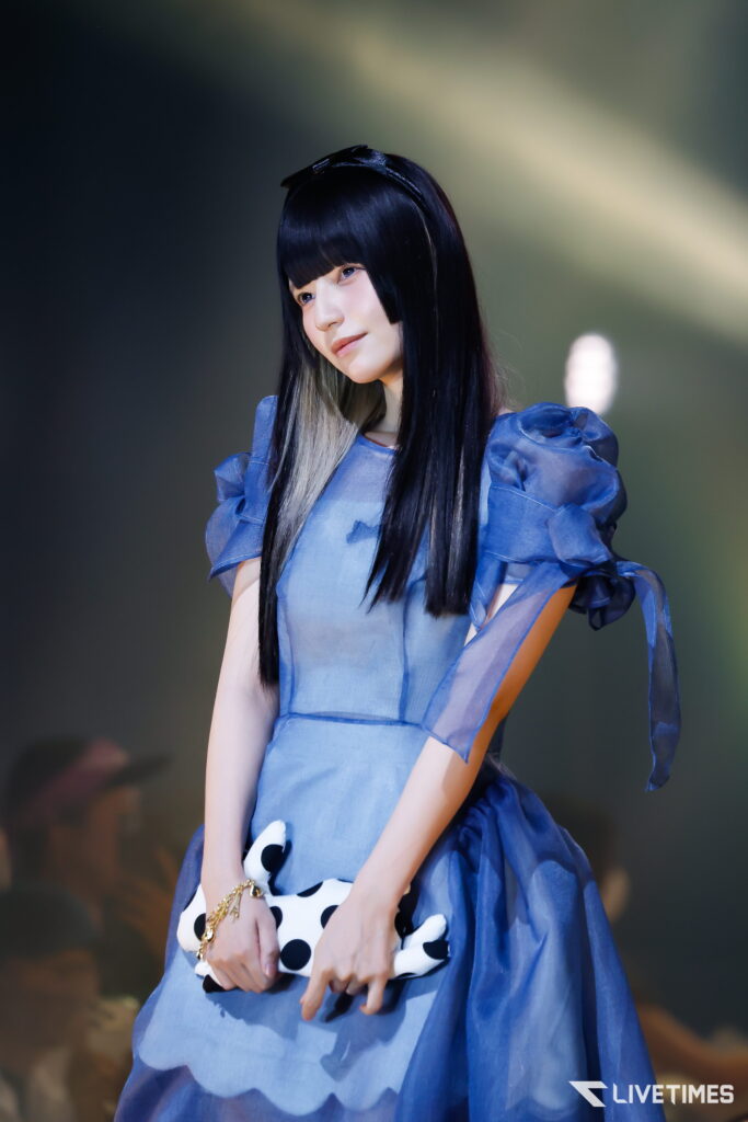 Young woman in a blue dress on stage, holding a white polka-dotted clutch with long black hair and a headband.