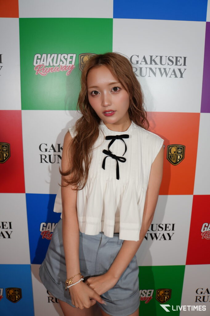Young woman with wavy brown hair in a white sleeveless top with black bow, posing in front of a colorful checkered backdrop with 'GAKUSEI RUNWAY' logos.