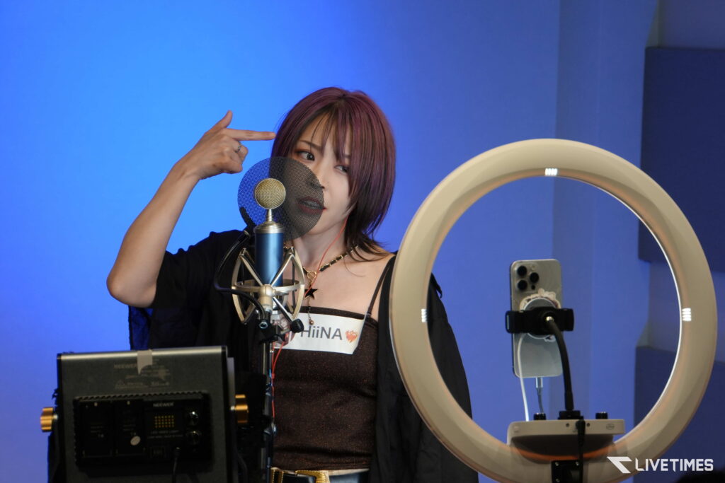 Female singer in a recording studio speaking/singing into a mic with a pop filter and ring light nearby on a blue backdrop.