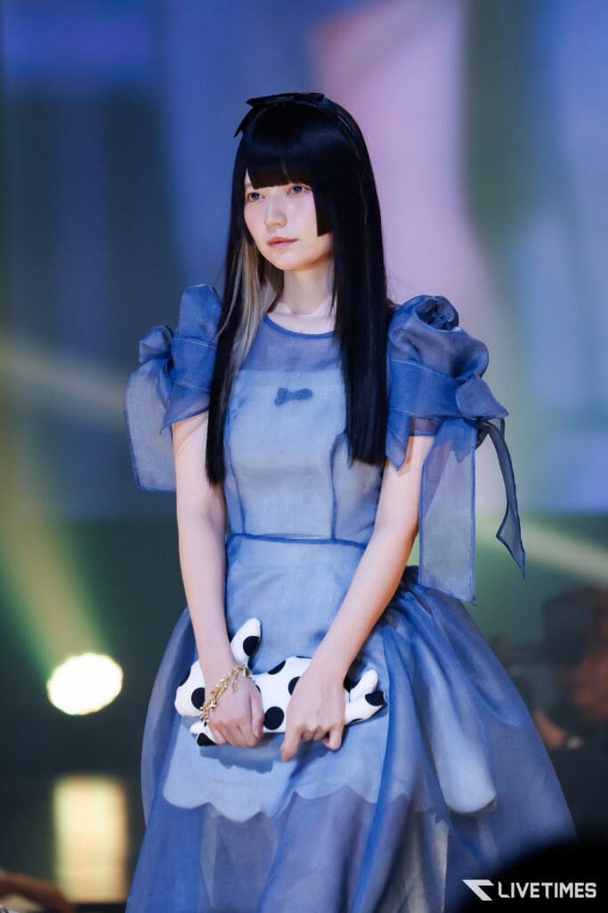 Young woman in a blue puff-sleeve dress on stage, holding a white polka-dot clutch with a calm expression.