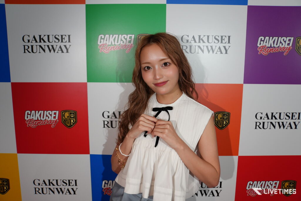 Young woman with wavy hair in a white sleeveless top posing in front of a colorful Gakusei Runway backdrop.