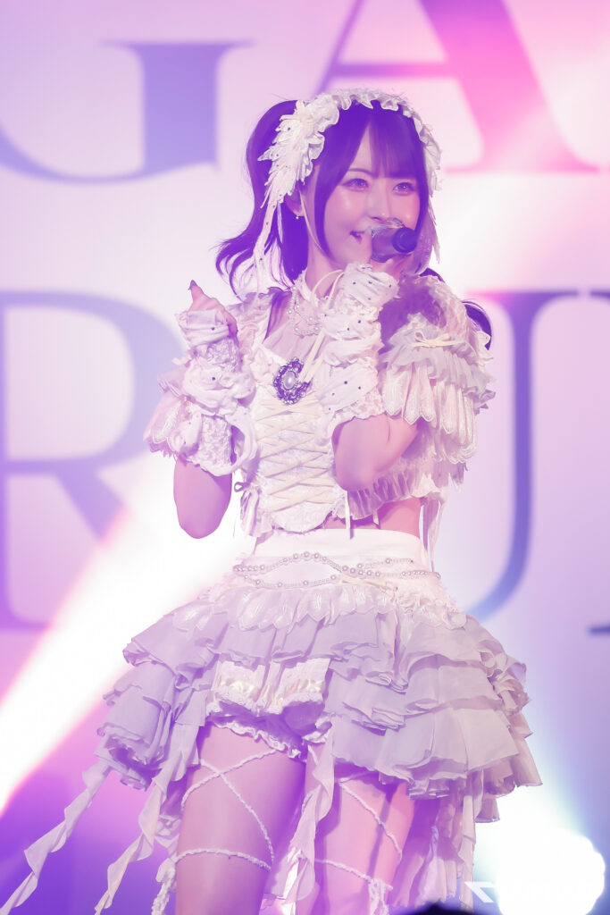 Female pop singer performing on stage in a white frilly dress with a flower headpiece, holding a microphone under pink-purple lights.