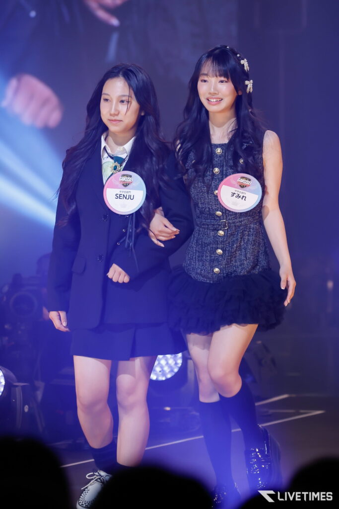 Two young performers walk arm-in-arm on a stage, wearing coordinated outfits and round event badges on their chests.