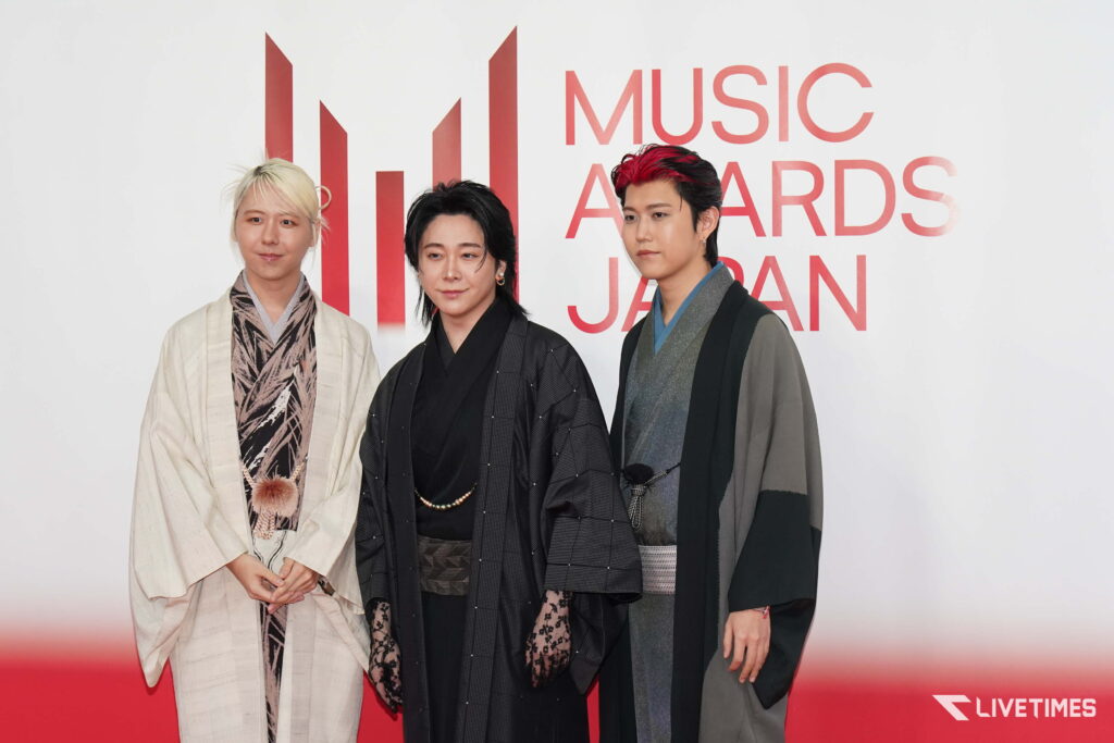 Three people in stylish, traditional-inspired outfits pose on a red carpet with a Music Awards Japan backdrop visible behind them.