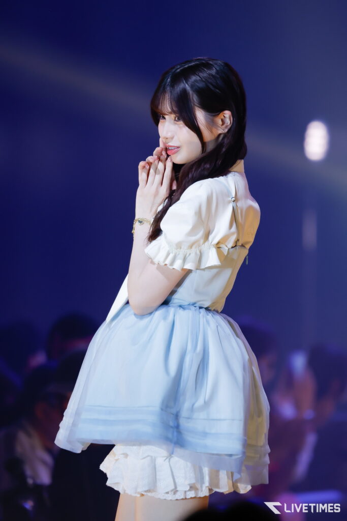 Female performer on stage, hands held near her face in a prayer-like pose, wearing a white blouse and light blue dress with a ruffled skirt, dark background.