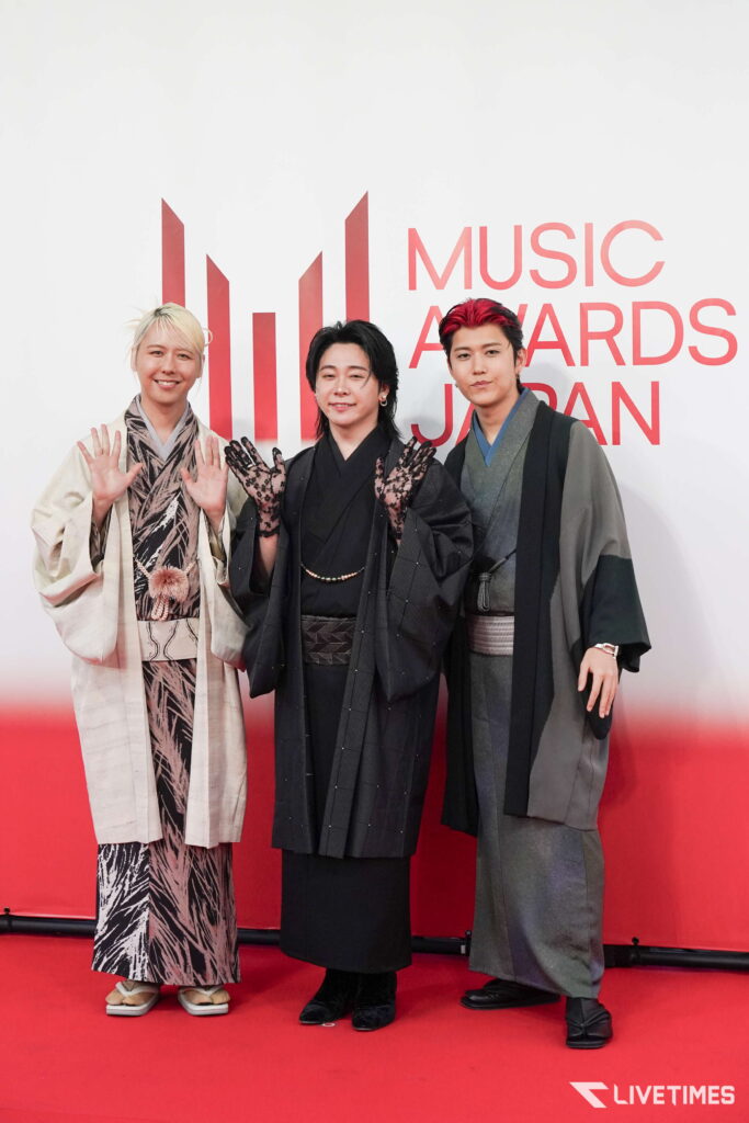 Three people in stylish kimono-style outfits pose on a red carpet, smiling and waving at the camera.