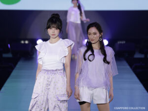 人気ライバー・早坂萌香&小夏みなみ、自然体のペアランウェイで視線集める 【アップティーサツコレ2026S/S】 Two models on a runway pose for the camera in pastel lavender and white outfits, with a blurred model visible in the background.