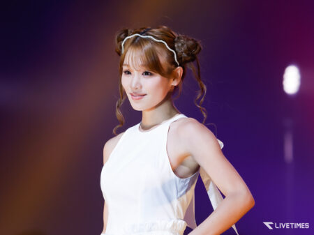 Portrait of a female performer on stage in a white sleeveless dress, hair in updo, soft stage lighting and a purple background.