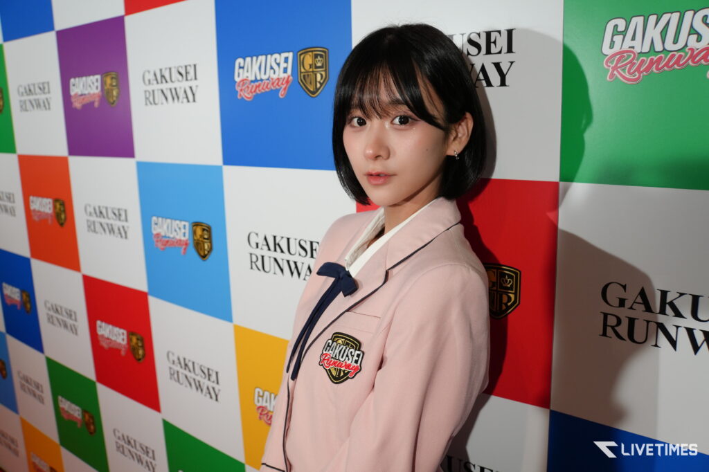 Model posing for photos at a Gakusei Runway event, wearing a pale pink blazer against a colorful sponsor backdrop.